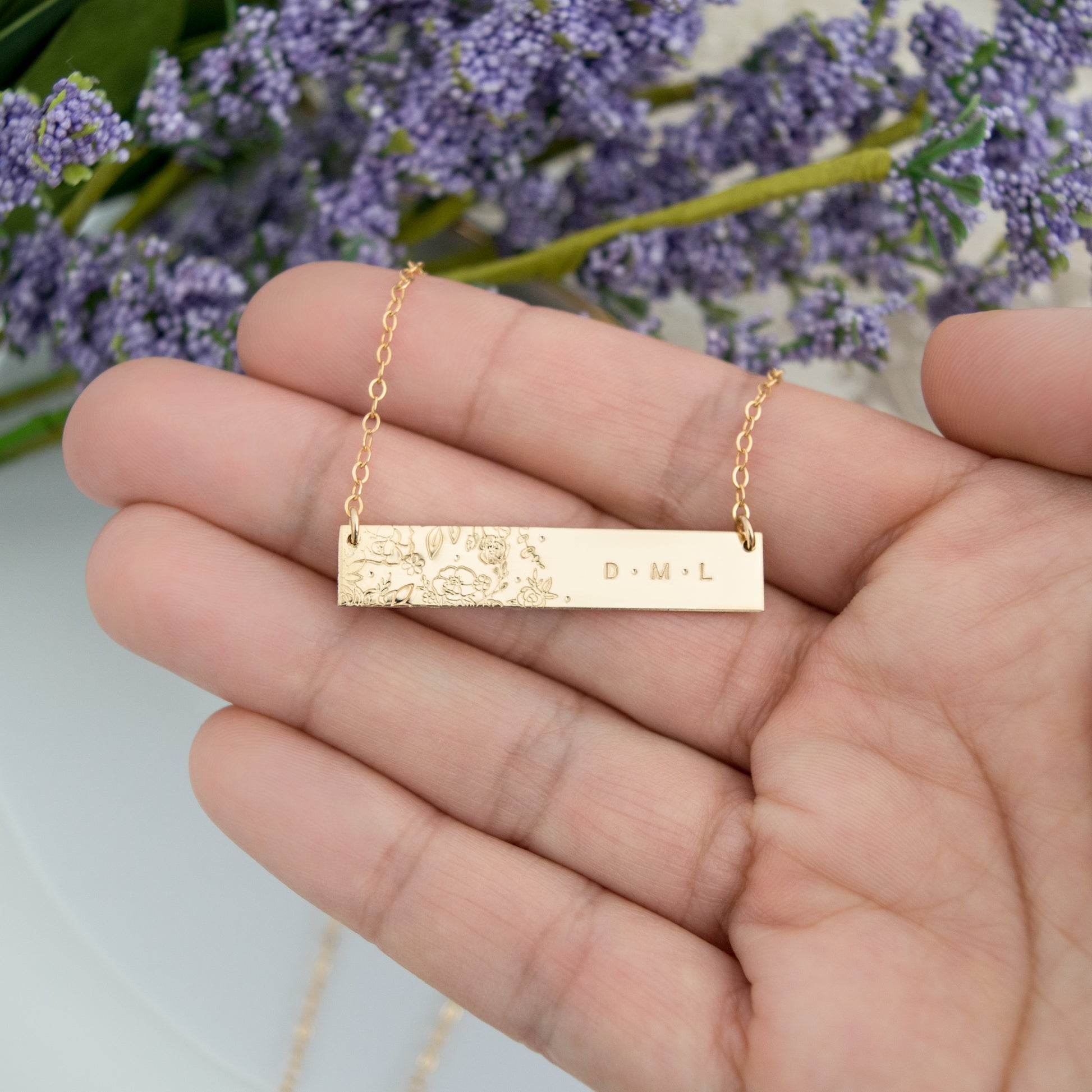 Hand holding the Wild Blooms Bar Necklace to show size and scale of the stamped floral and personalized details.